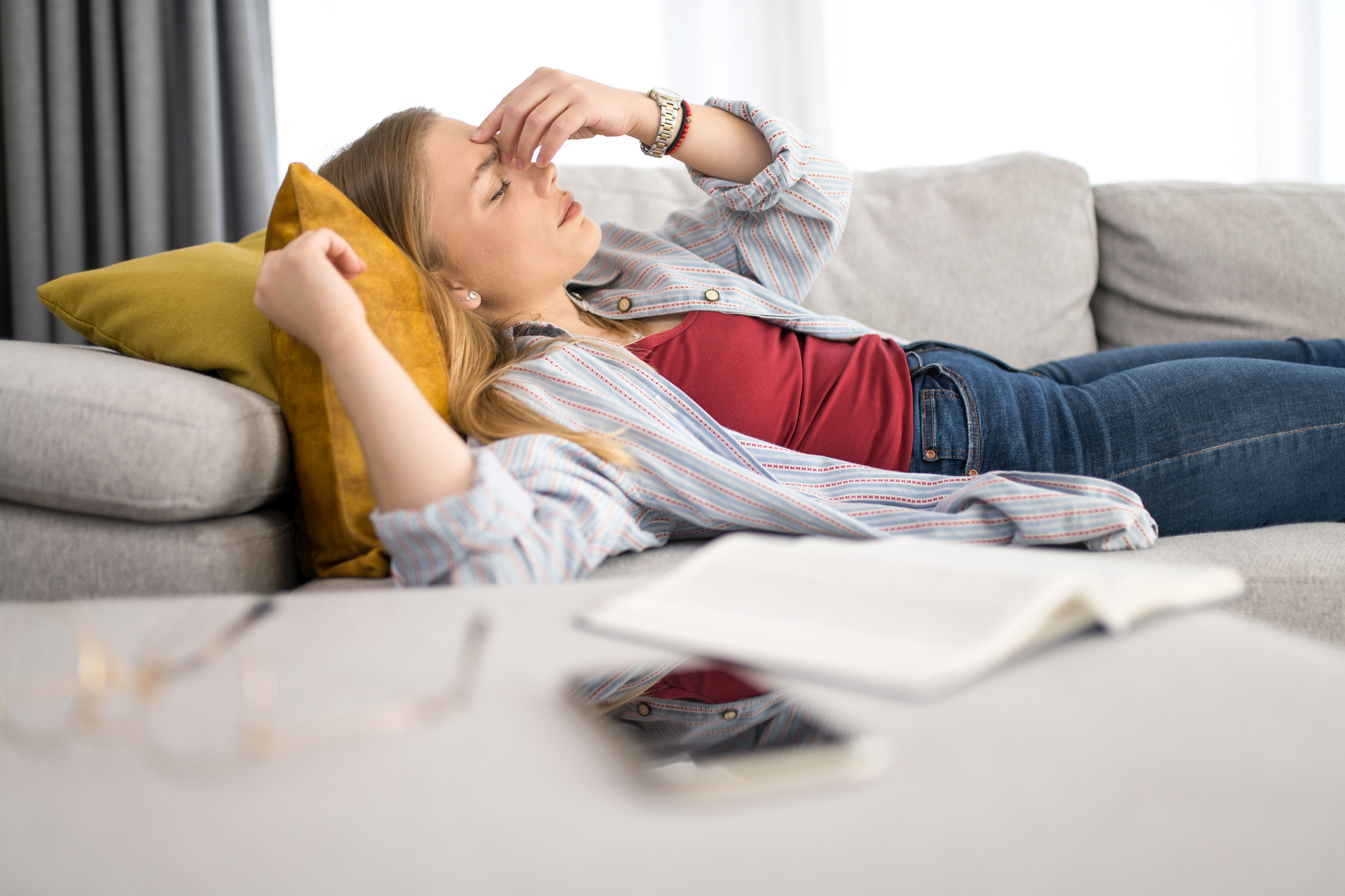 A young woman lying on a sofa, holding her forehead in a gesture of pain or stress, with an open book and glasses in the foreground.